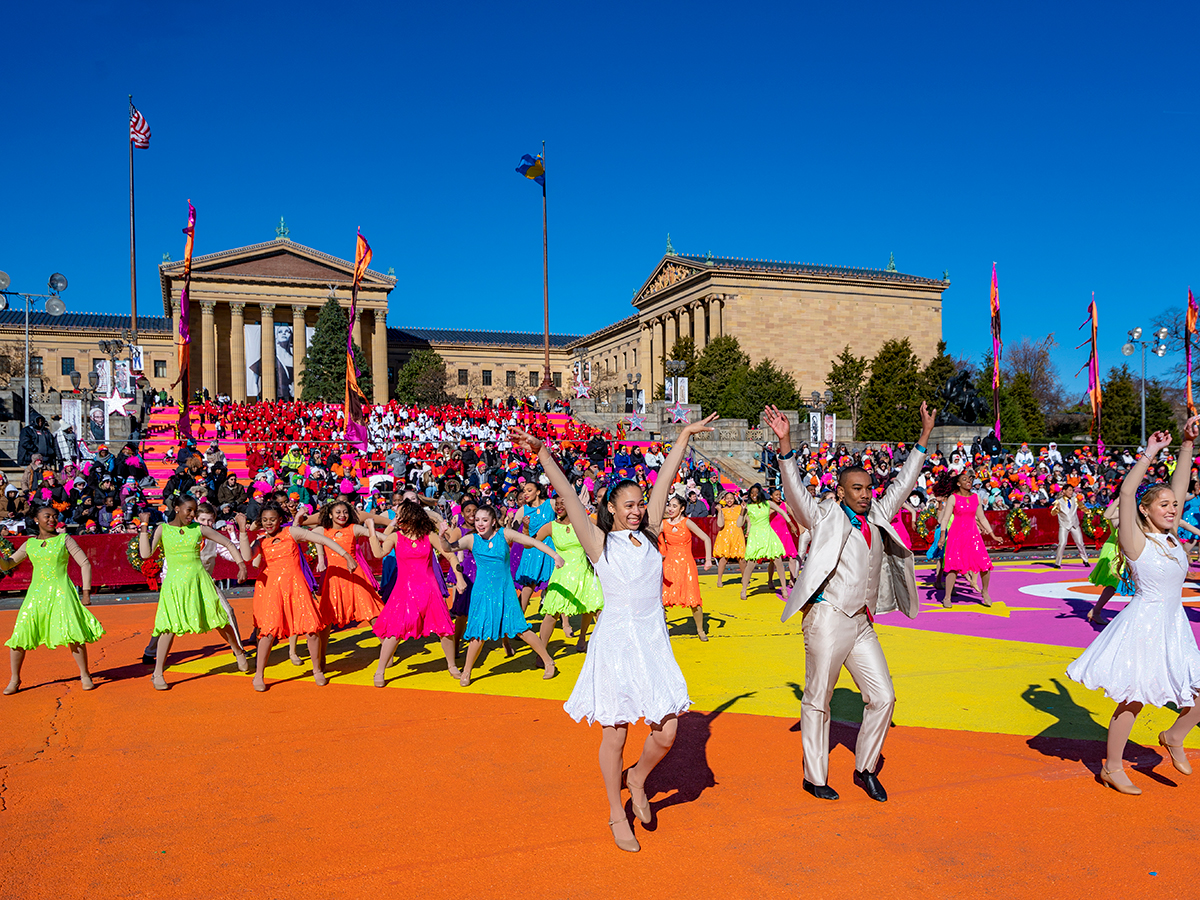 Performers in bright, colorful costumes dance in front of the Philadelphia Art Museum during the annual Thanksgiving Day parade, while a crowd watches from the Art Museum steps.