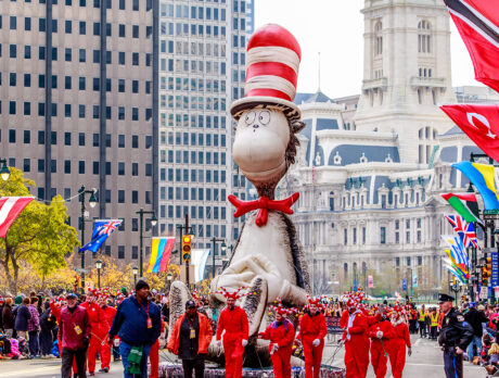 A large Cat in the Hat float being marched down the Benjamin Franklin Parkway during the Thanksgiving Parade in Philadelphia, with City Hall in the background and crowds of people watching from the sidewalks.