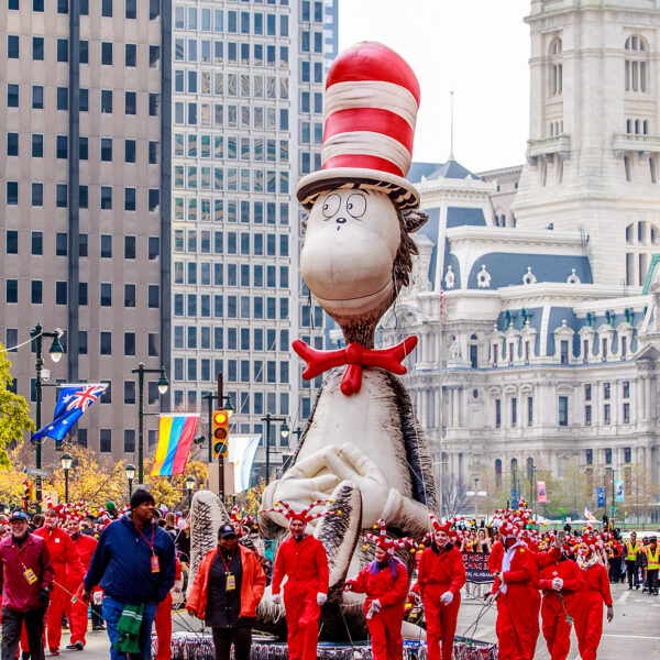 A large Cat in the Hat float being marched down the Benjamin Franklin Parkway during the Thanksgiving Parade in Philadelphia, with City Hall in the background and crowds of people watching from the sidewalks.