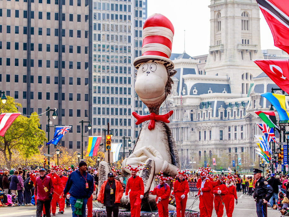 A large Cat in the Hat float being marched down the Benjamin Franklin Parkway during the Thanksgiving Parade in Philadelphia, with City Hall in the background and crowds of people watching from the sidewalks.