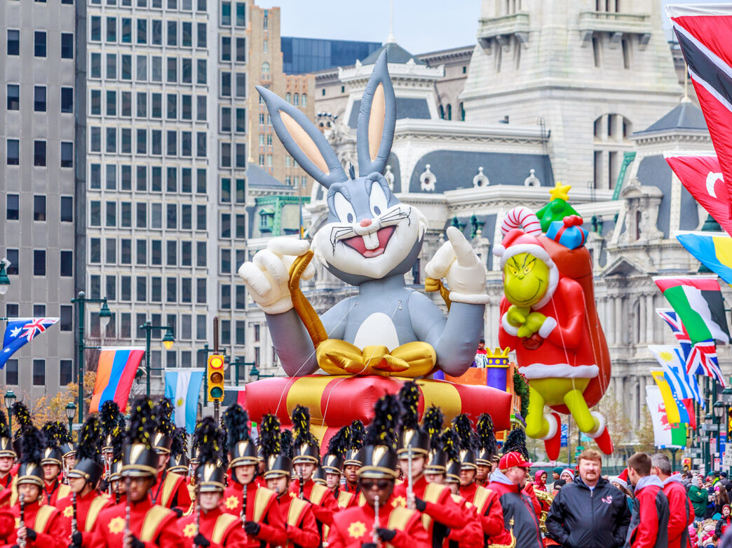 Large inflatable balloon floats of Bugs Bunny and The Grinch float above a marching bands and crowds along the Benjamin Franklin Parkway with Philadelphia's City Hall in the background.