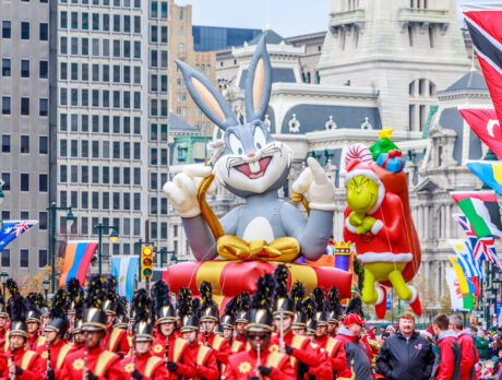 Large inflatable balloon floats of Bugs Bunny and The Grinch float above a marching bands and crowds along the Benjamin Franklin Parkway with Philadelphia's City Hall in the background.