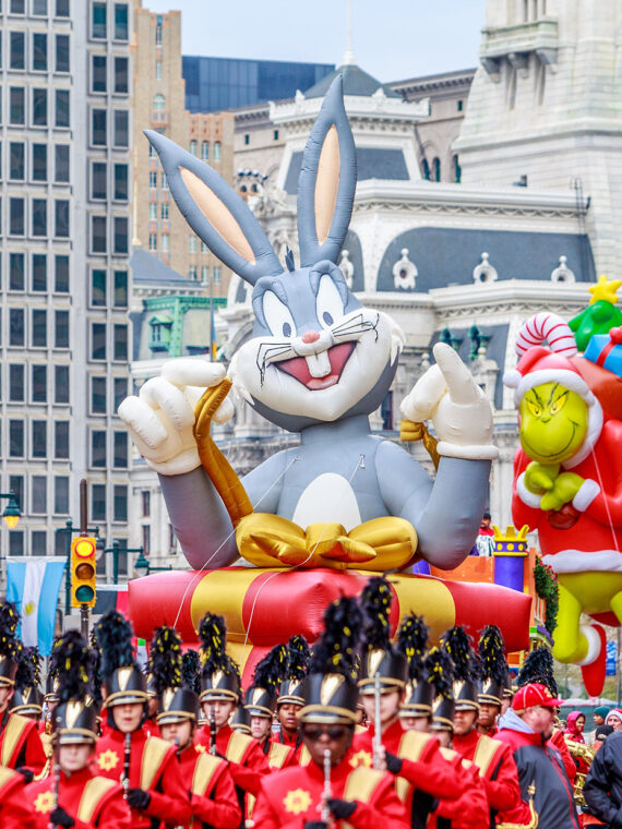 Large inflatable balloon floats of Bugs Bunny and The Grinch float above a marching bands and crowds along the Benjamin Franklin Parkway with Philadelphia's City Hall in the background.