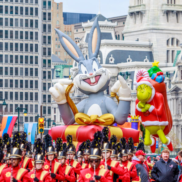 Large inflatable balloon floats of Bugs Bunny and The Grinch float above a marching bands and crowds along the Benjamin Franklin Parkway with Philadelphia's City Hall in the background.
