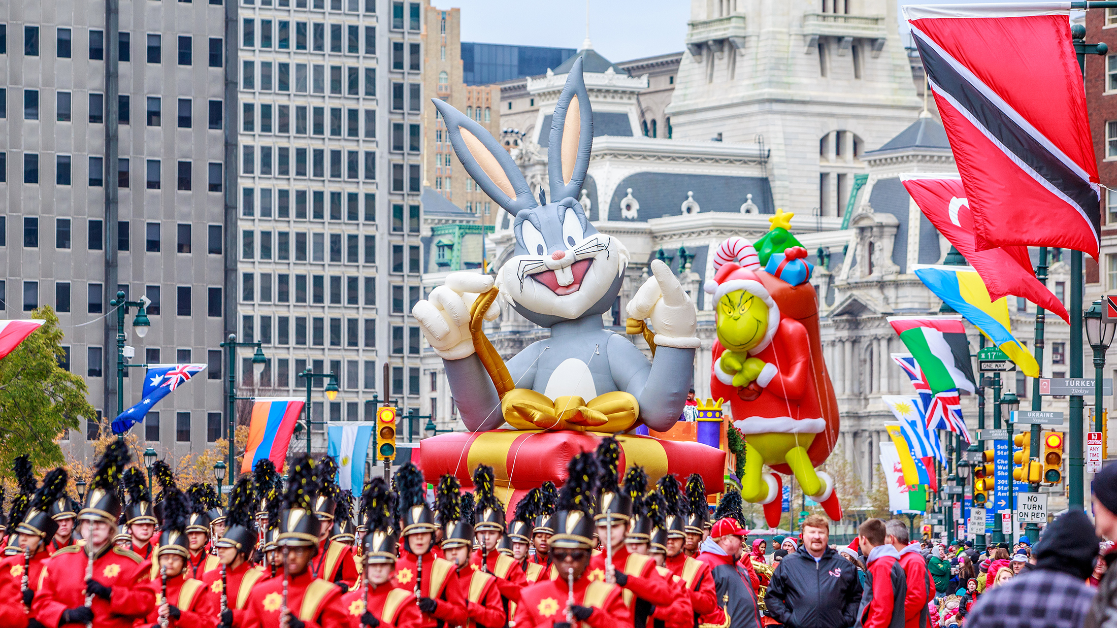 Large inflatable balloon floats of Bugs Bunny and The Grinch float above a marching bands and crowds along the Benjamin Franklin Parkway with Philadelphia's City Hall in the background.