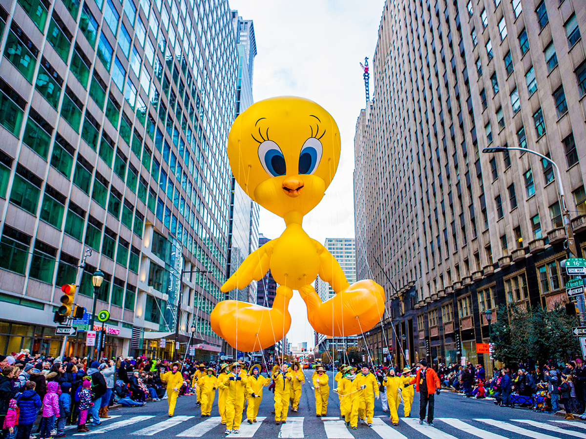 Parade participants in yellow jumpsuits guide a massive Tweety Bird balloon float through Center City streets lined with spectators and tall buildings.