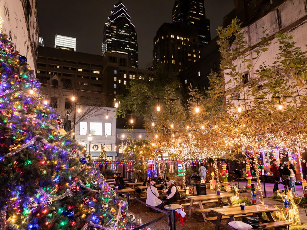 A festive outdoor beer gardens decorated with string lights, picnic tables and a large Christmas tree with multicolored lights, with city skyscrapers in the background at night.