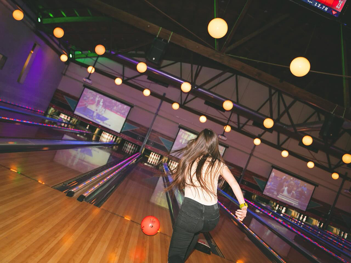 A woman rolls a red bowling ball along a bowling lane, with sports games playing on big screens in the background.