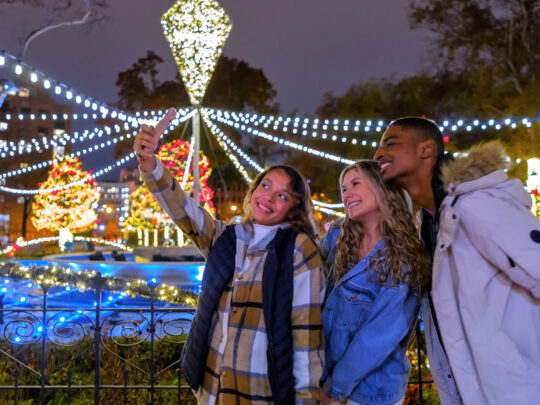 Three young adults wearing jackets and scarfs pose for a photo in front of a display of string lights at Franklin Square.