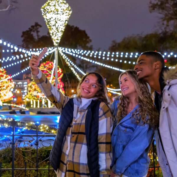 Three young adults wearing jackets and scarfs pose for a photo in front of a display of string lights at Franklin Square.