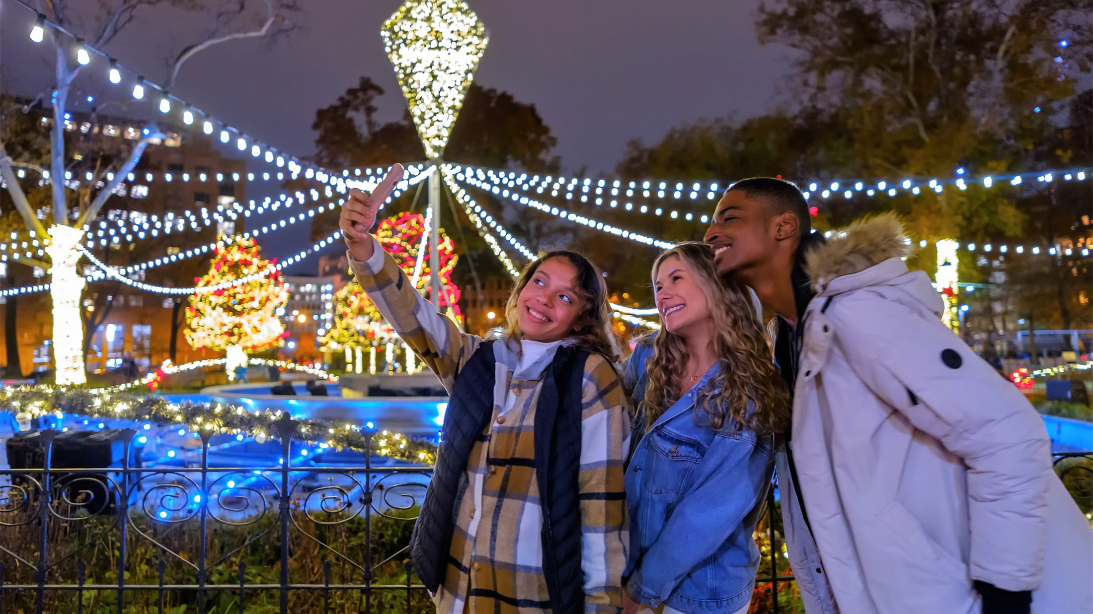 Three young adults wearing jackets and scarfs pose for a photo in front of a display of string lights at Franklin Square.