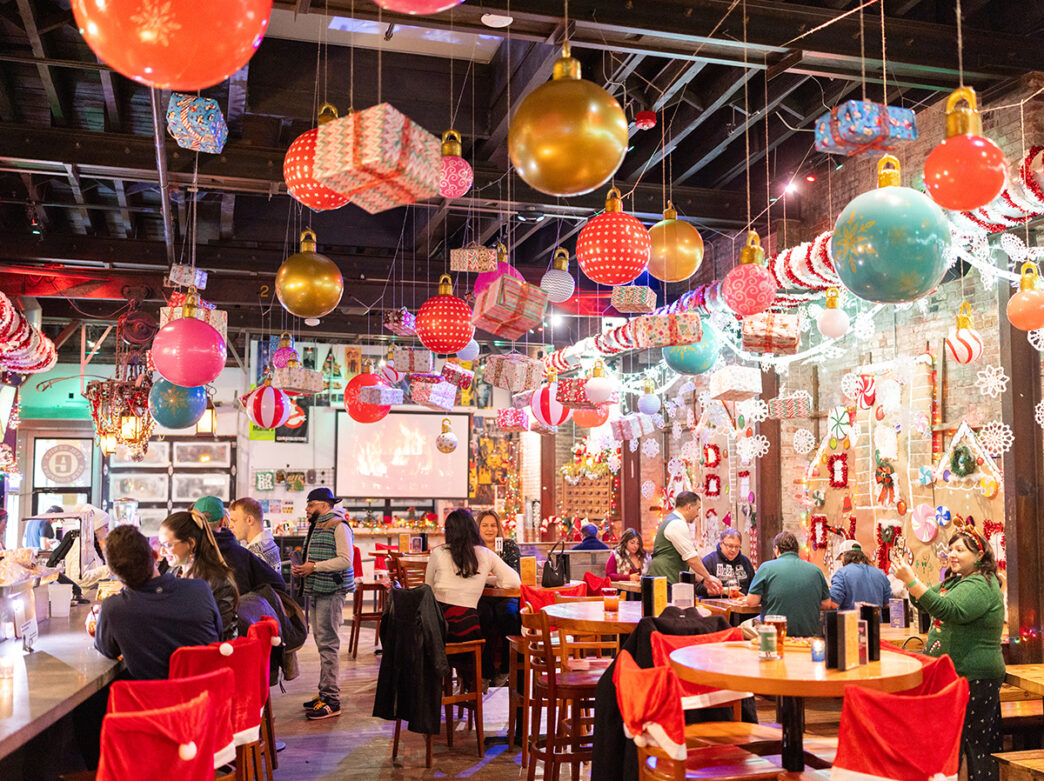 The inside of a holiday-themed bar filled with oversized ornaments and wrapped gift boxes hanging from the ceiling as diners enjoy food and drinks.