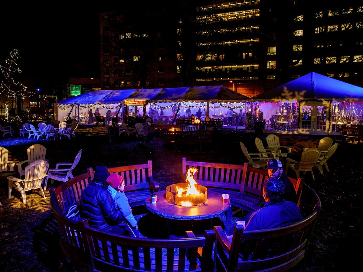 People sit around a glowing fire pit at night in Franklin Square, surrounded by twinkling lights, outdoor seating and a festively lit tented area.