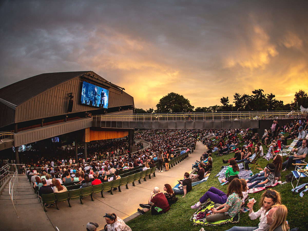 Crowds of people relax on the lawn and under the pavilion seating at the Mann Center during and outdoor concert, as a glowing sunset fills the sky above the stage.