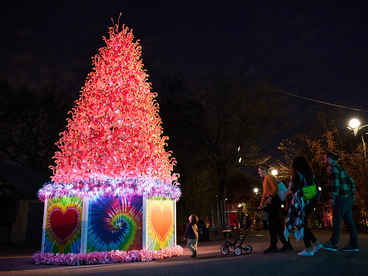 A family with a child and stroller admires a towering pink tree made entirely of illuminated flamingo figures at night.