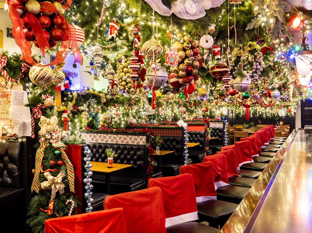 A restaurant interior is lavishly decorated for the holidays, with dense arrangements of twinkling lights, ornaments, garlands and Santa hat chair covers creating an over the top festive atmosphere.