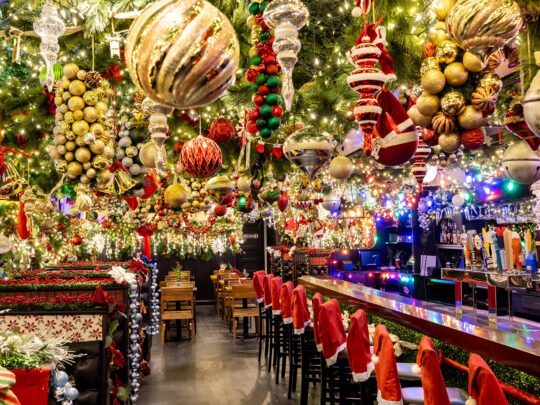 A restaurant interior overflowing with holiday decor, including oversize ornaments, garland and twinkling lights hanging from the ceiling. Plus, Santa hate chair covers over the backs of barstools.