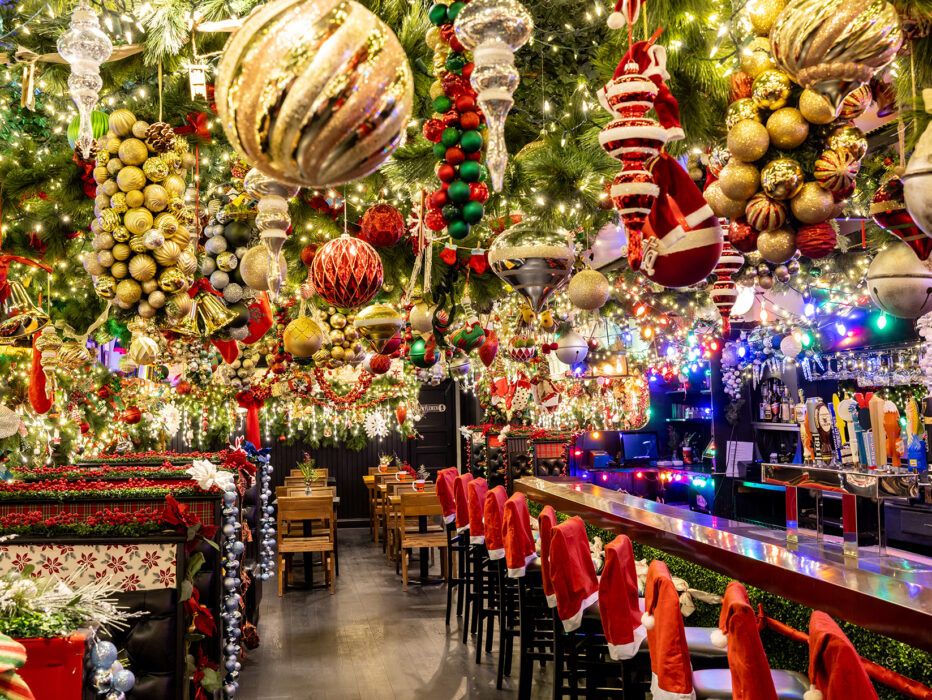 A restaurant interior overflowing with holiday decor, including oversize ornaments, garland and twinkling lights hanging from the ceiling. Plus, Santa hate chair covers over the backs of barstools.