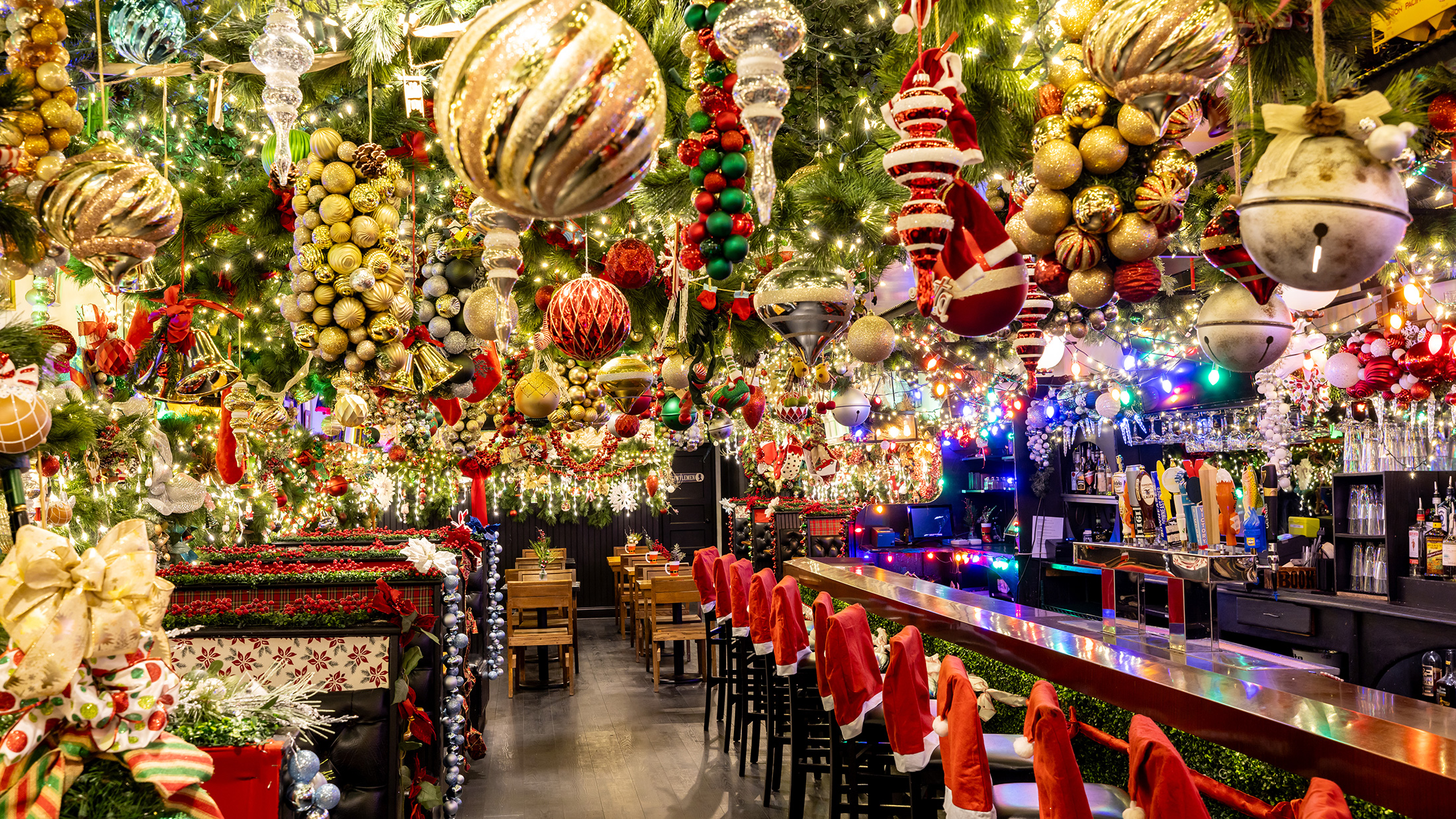 A restaurant interior overflowing with holiday decor, including oversize ornaments, garland and twinkling lights hanging from the ceiling. Plus, Santa hate chair covers over the backs of barstools.