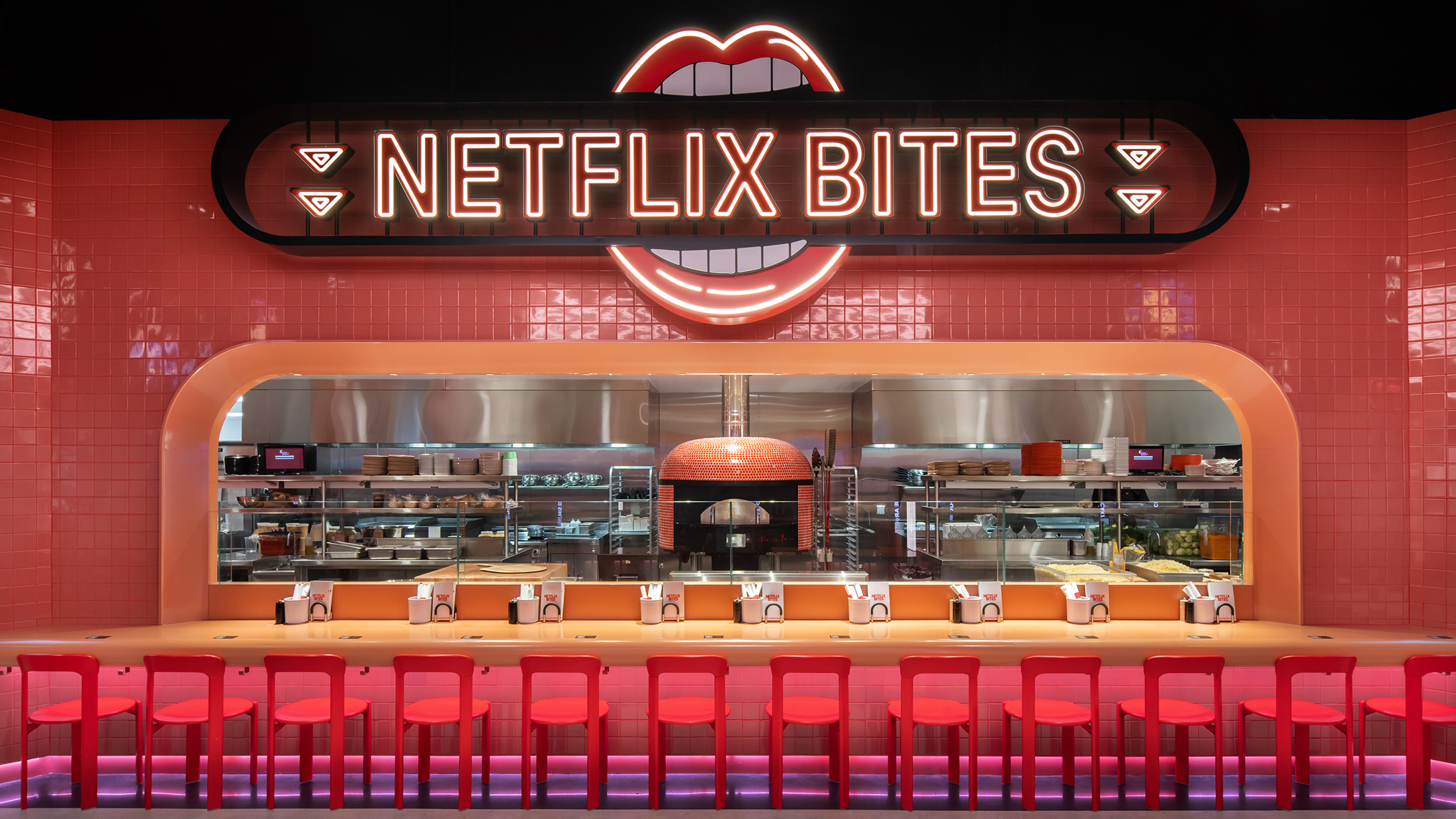 A bright, neon-lit food counter with red stools lined in front of an open kitchen featuring a pizza oven, framed by glossy red tile and a glowing sign shaped like lips above.