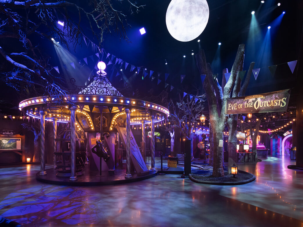 A dim, moody carnival scene featuring a glowing carousel, surrounded by eerie trees, hanging pennants and dramatic blue lighting beneath a large full moon.