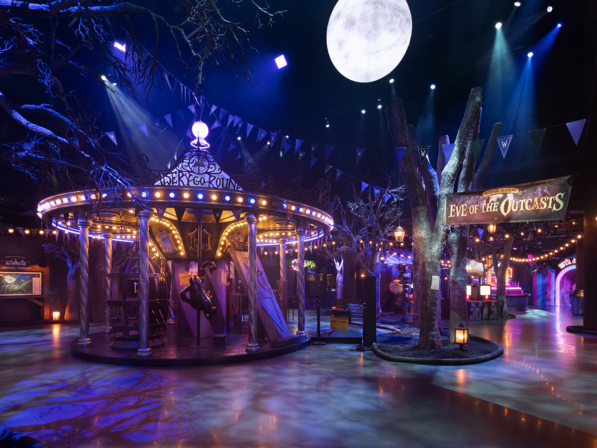 A dim, moody carnival scene featuring a glowing carousel, surrounded by eerie trees, hanging pennants and dramatic blue lighting beneath a large full moon.