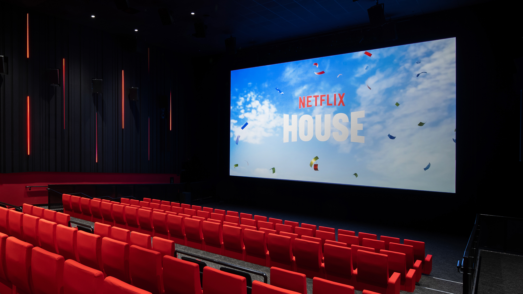 A modern theater with rows of bright red seats facing a massive screen displaying the Netflix House logo against a blue sky backdrop.
