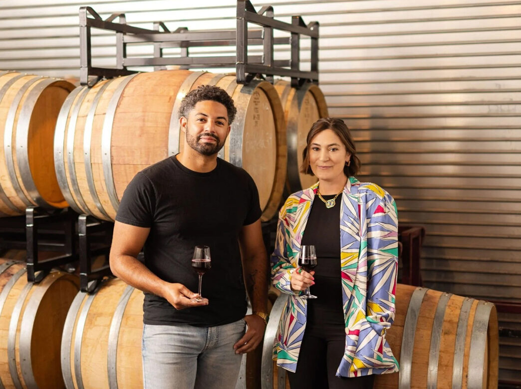 A man and a woman stand in front of stacked wooden wine barrels, each holding a glass of red wine and smiling at the camera.