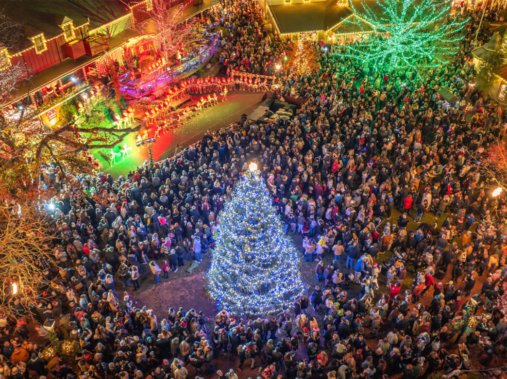 An aerial view of a crowd of visitors gathered around a glowing Christmas tree at Peddler's Village during the Grand Illumination celebration.