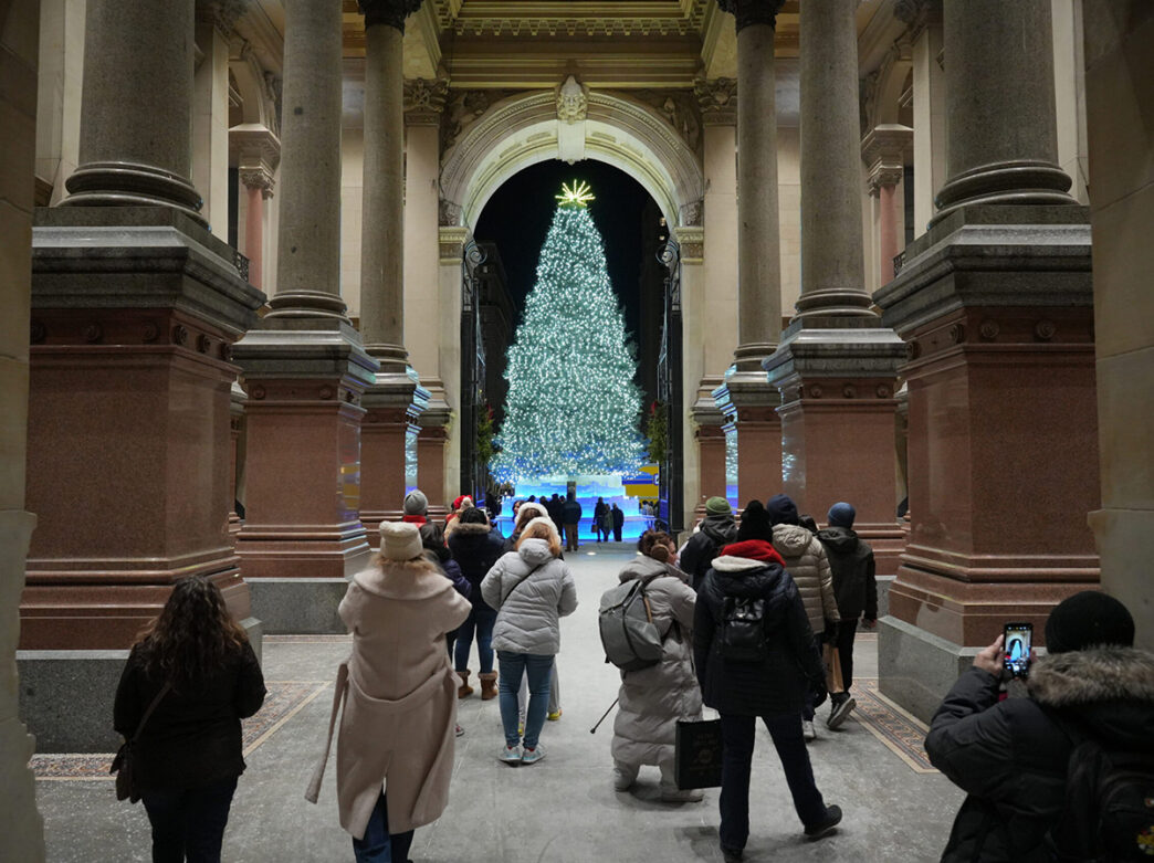 Visitors bundled in winter coats walk through a grand archway of Philadelphia's City Hall toward a massive Christmas tree covered in white lights, framed by tall stone columns.