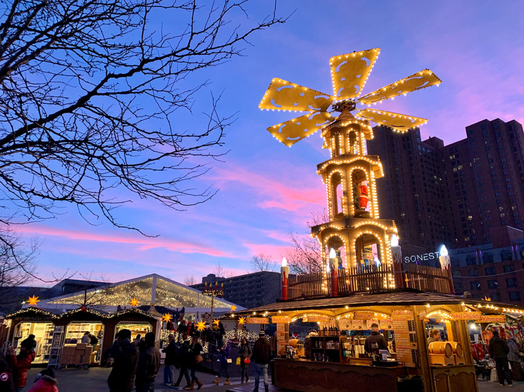 A large illuminated Christmas pyramid towers over festival stalls at a Christmas village as visitors shop and explore. Warm lighting and holiday decor glow against the pink and blue sunset sky.