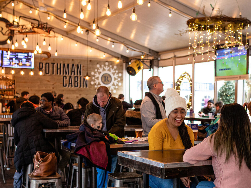 People gather inside a temporary cabin decorated with string lights and ornament chandeliers, chatting over drinks.