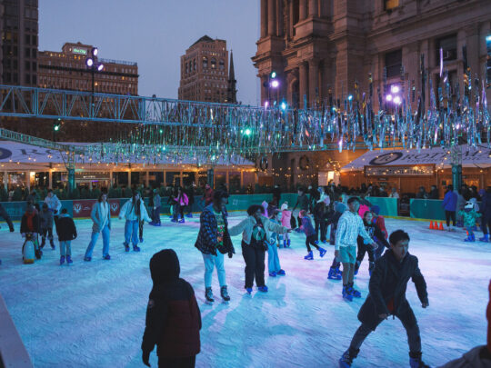 Crowds of skaters enjoy an evening on the Rothman Orthopaedics Ice Rink at Dilworth Park, with people gliding across the ice under sparkling silver decorations.