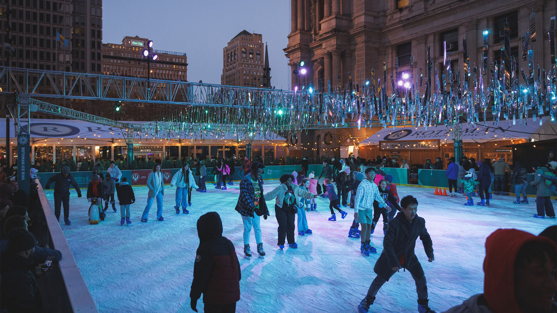 Crowds of skaters enjoy an evening on the Rothman Orthopaedics Ice Rink at Dilworth Park, with people gliding across the ice under sparkling silver decorations.