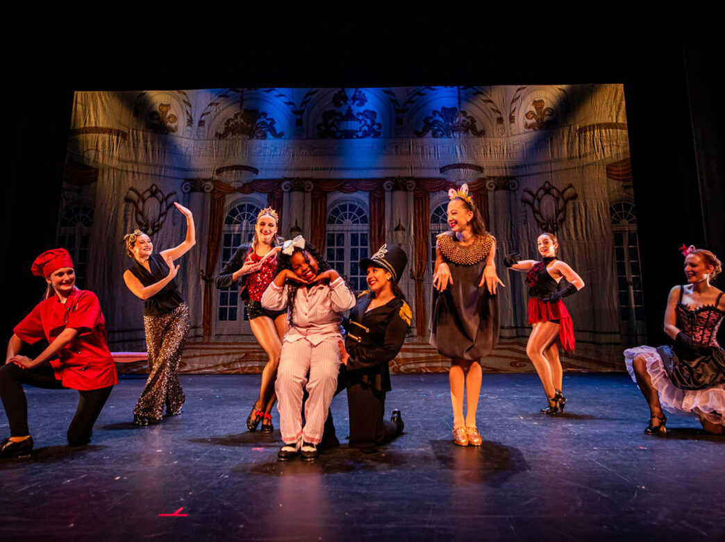 A group of tap dancers dresses in costumes strike playful poses on stage, one in pajamas at the center sitting on the bent knee of a dancer dresses as a soldier.