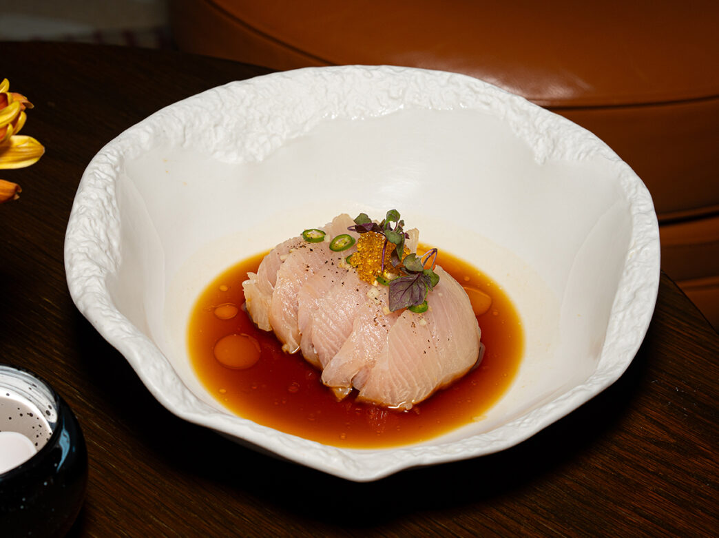 A delicate serving of sashimi-style white fish sits in a shallow soy-based broth, topped with microgreens and thin chili slices.
