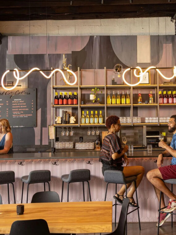 Guests chat and sip wine at a bar inside a modern, cozy wine tasting room, with shelves of colorful wine bottles and soft, curved pendant lights overhead.