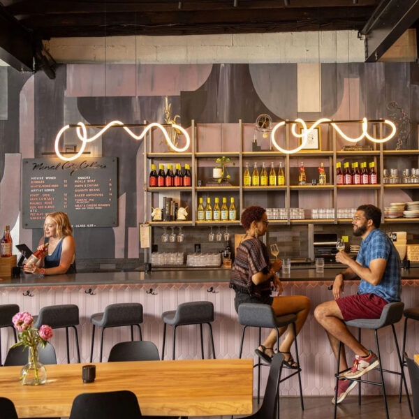 Guests chat and sip wine at a bar inside a modern, cozy wine tasting room, with shelves of colorful wine bottles and soft, curved pendant lights overhead.