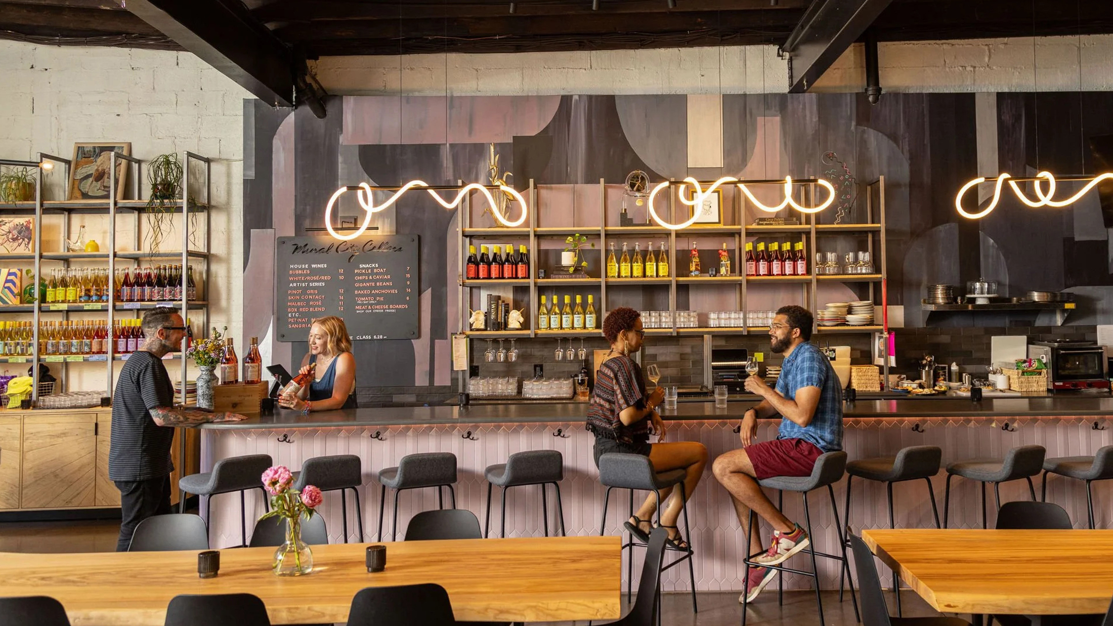 Guests chat and sip wine at a bar inside a modern, cozy wine tasting room, with shelves of colorful wine bottles and soft, curved pendant lights overhead.