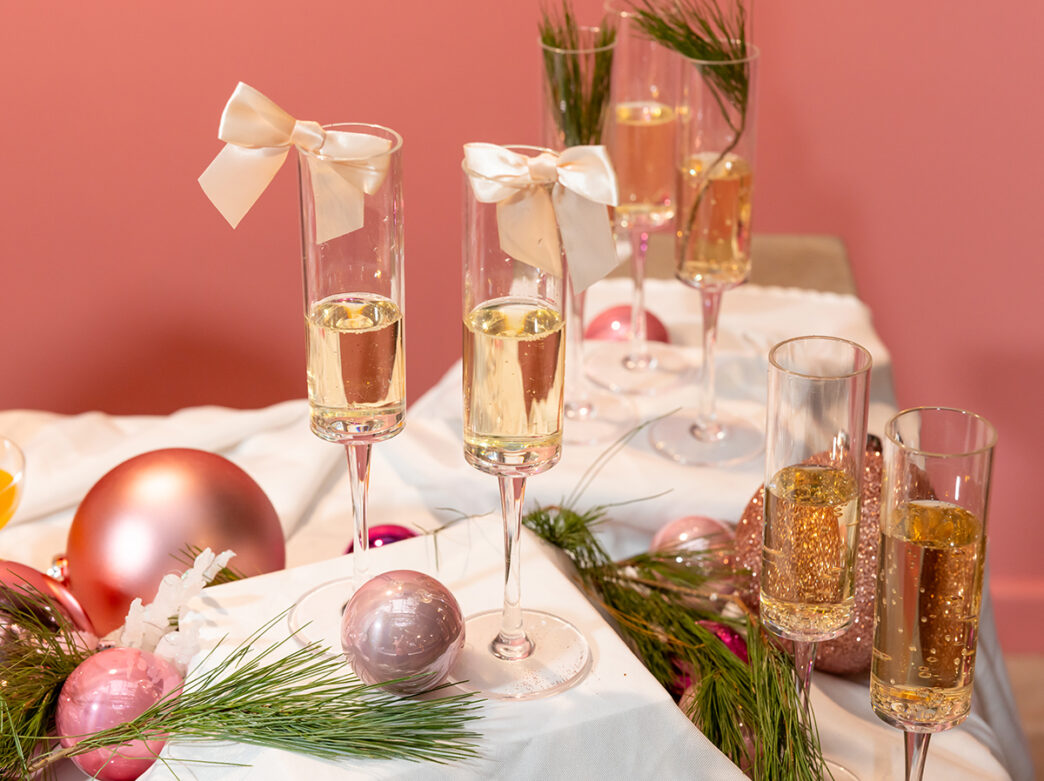 Champagne flutes topped with silk white bows arranged on a white cloth, surrounded by pink ornaments and evergreen sprigs against a soft pink backdrop.
