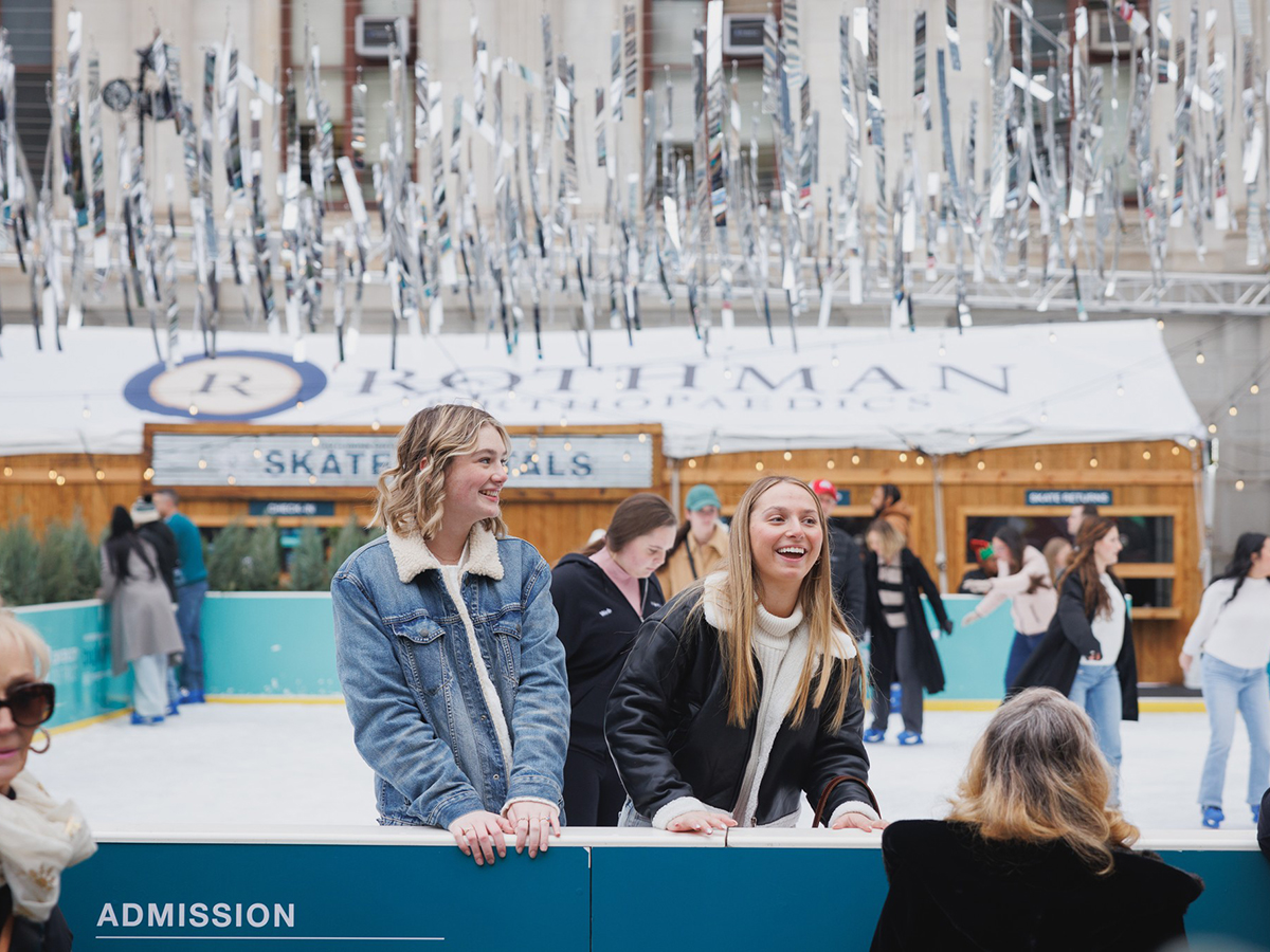 Two people smile and lean against the barrier at the Rothman Orthopaedics Ice Rink as skaters glide across the ice under sparkling silver decorations.