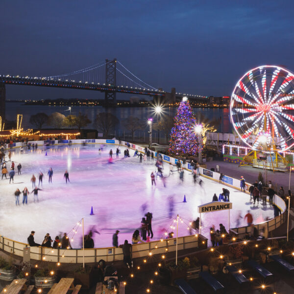 Skaters glide across an ice rink with a Ferris wheel and holiday tree sparkle against the Delaware River and Benjamin Franklin Bridge in the background.