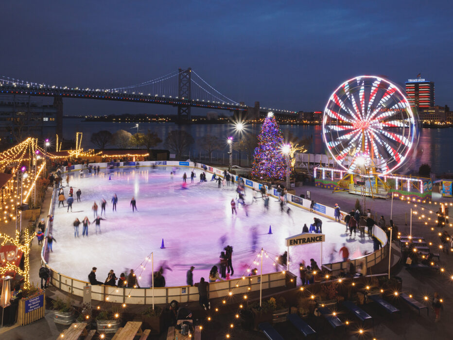 Skaters glide across an ice rink with a Ferris wheel and holiday tree sparkle against the Delaware River and Benjamin Franklin Bridge in the background.