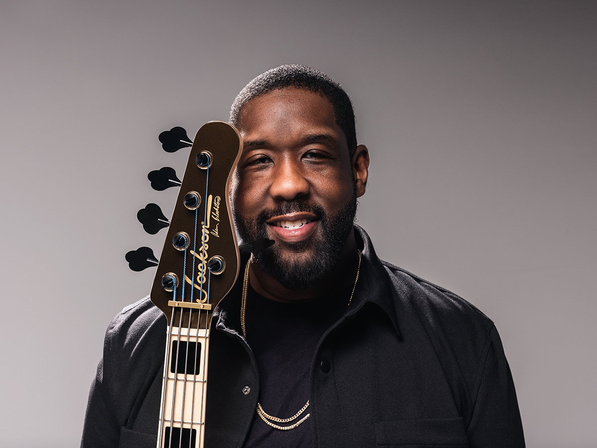 A musician smiles at the camera while holding a base guitar upright against his shoulder, wearing a black jacket and golden chain against a neutral studio backdrop.