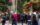 A crowd gathers in a decorated Old City street for a Hanukkah celebration, with a large menorah and American flag standing beneath tall trees. People in winter coats chat and stroll past festive garlands hung with red ornaments.