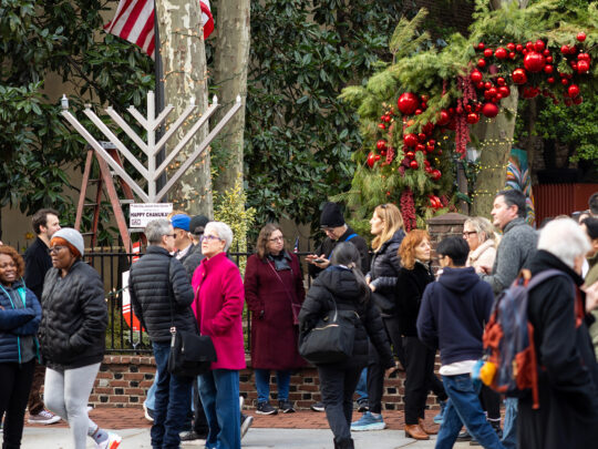 A crowd gathers in a decorated Old City street for a Hanukkah celebration, with a large menorah and American flag standing beneath tall trees. People in winter coats chat and stroll past festive garlands hung with red ornaments.