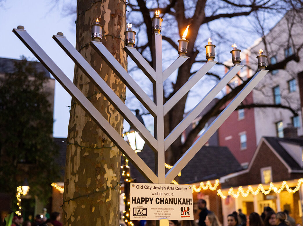 A large outdoor menorah with several candles lit in front of a tree, surrounded by holiday lights and Old City buildings at dusk.