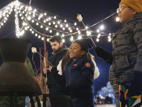 Children wearing winter jackets, hats and gloves roast marshmallows over a fire pit under a canopy of string lights.