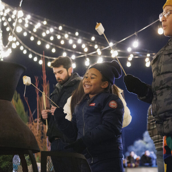Children wearing winter jackets, hats and gloves roast marshmallows over a fire pit under a canopy of string lights.