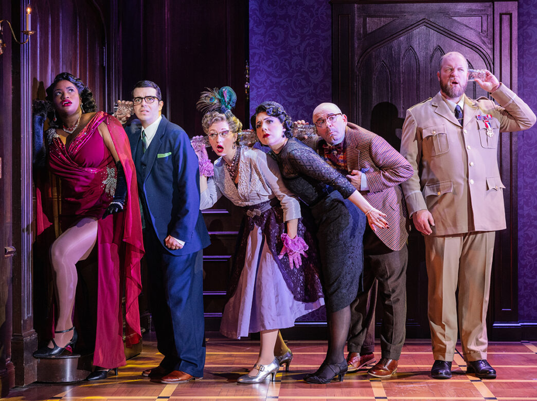 Des acteurs en costumes d'époque se penchent en tenant des coupes en verre sur leur tête pour essayer d'écouter la pièce d'à côté pendant la production de Clue.